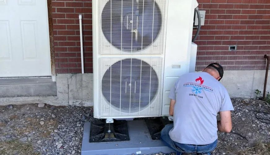 Technician kneels beside an outdoor HVAC unit against a brick wall, inspecting equipment.