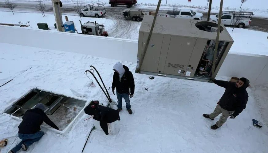 People on a snowy rooftop near an open hatch and large HVAC unit, one holding cables.
