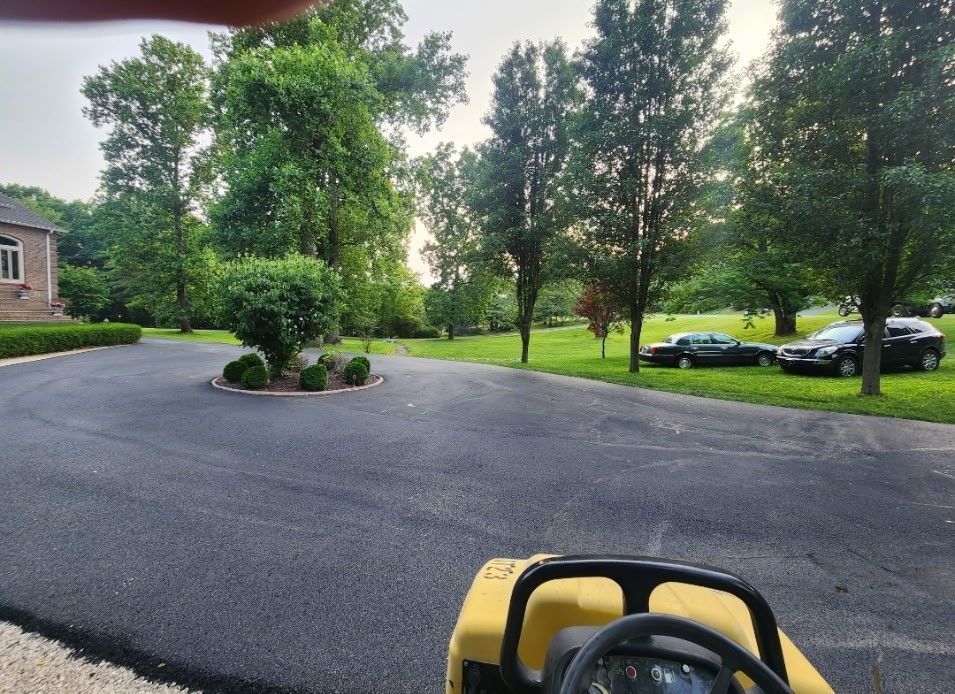 Asphalt driveway with a small island of bushes; trees and cars in the background