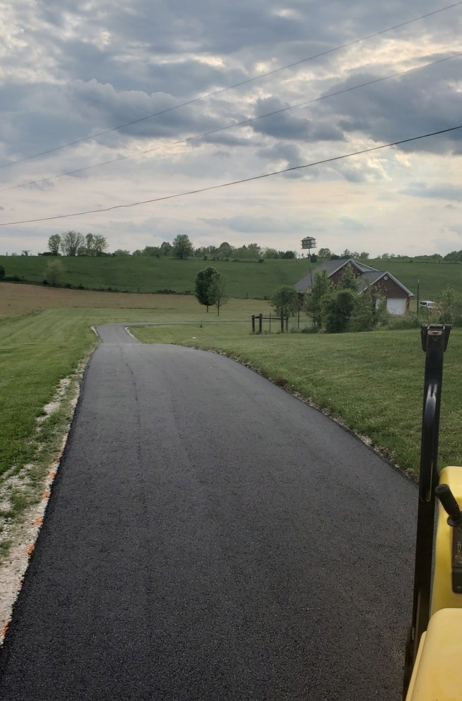 Asphalt driveway winding towards a house, surrounded by green grass and rolling hills under a cloudy sky