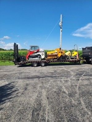 A trailer loaded with construction equipment, including a skid steer, parked outdoors under a blue sky
