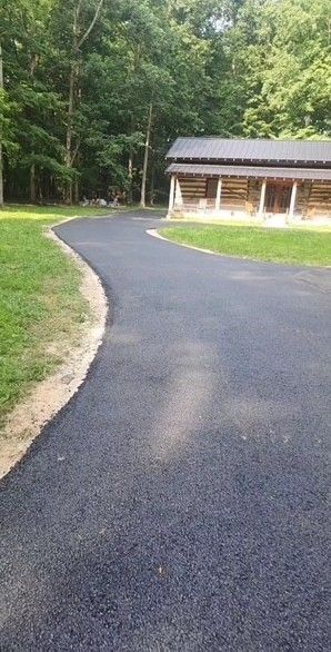 Paved asphalt driveway curves toward a wooden log building; green grass and trees surround