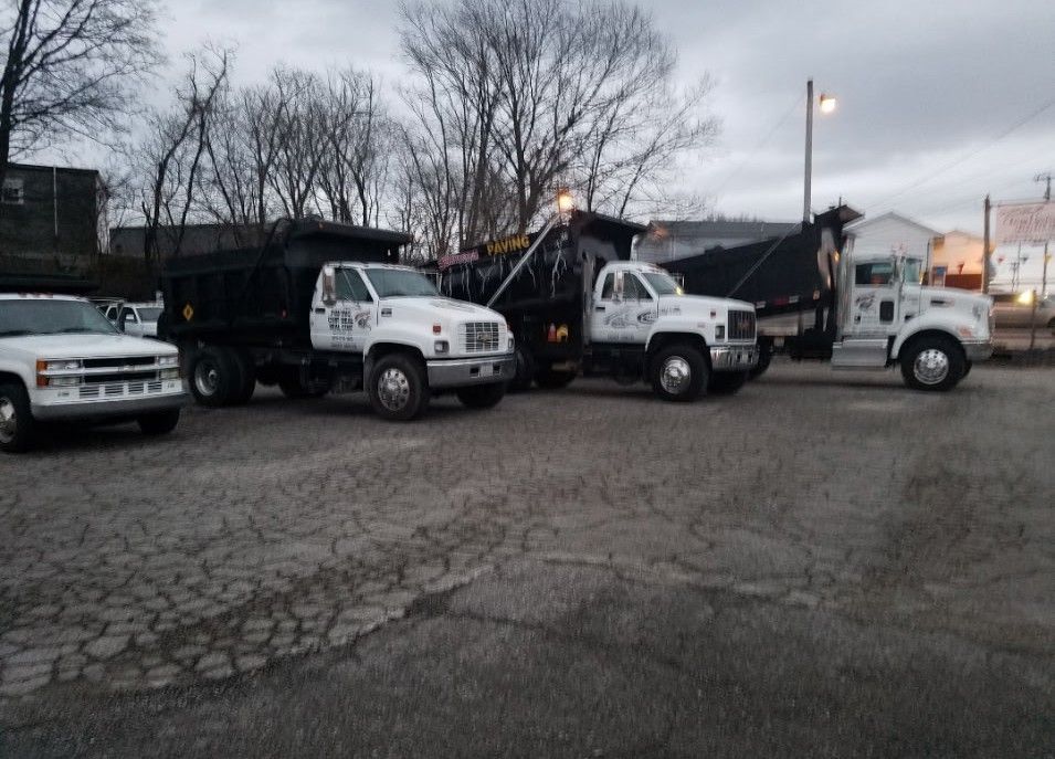 Four white dump trucks parked on a cracked asphalt lot, under a cloudy sky