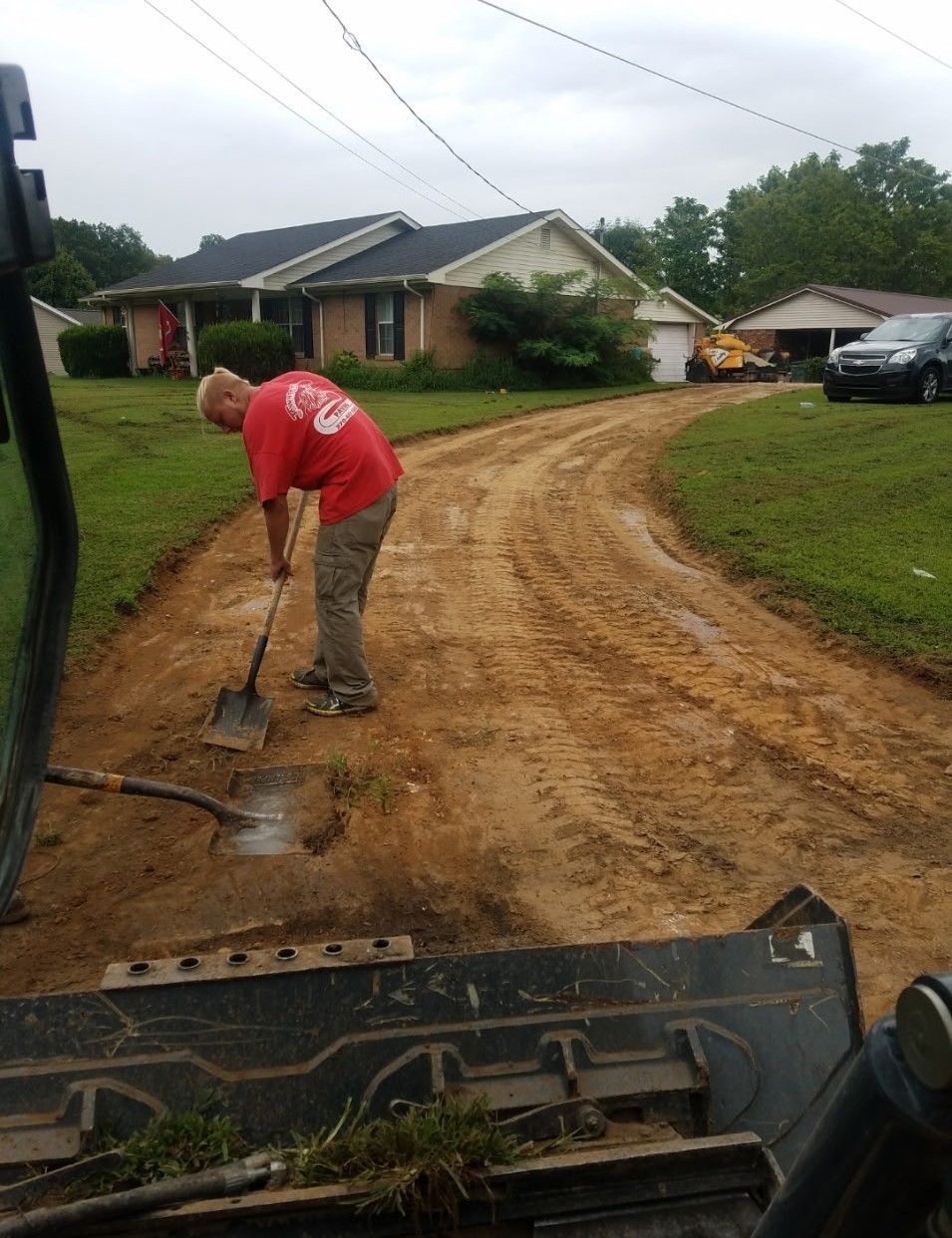 Man shoveling dirt on a muddy driveway, with a house and overcast sky in the background