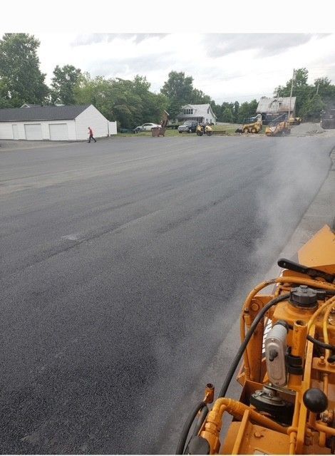 Newly paved asphalt surface with construction equipment; a worker stands in the distance
