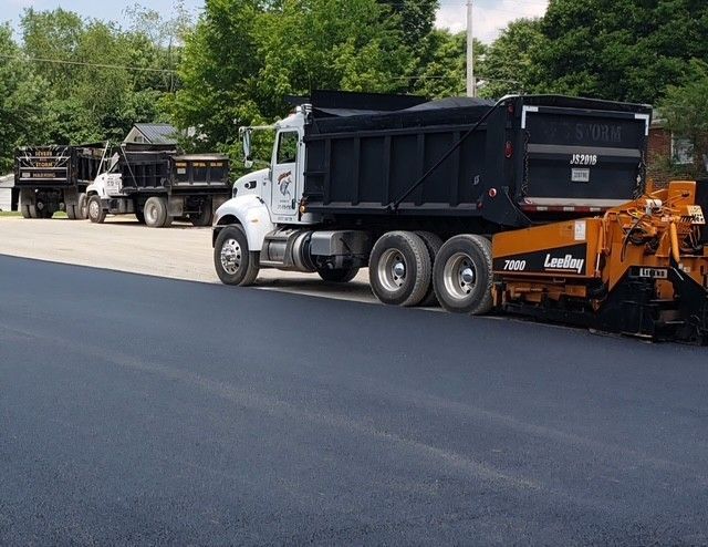 Asphalt paving in progress: dump trucks next to paving machine on dark asphalt, trees in background