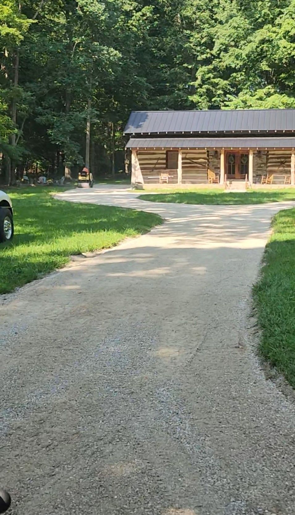 Gravel driveway leads to a log cabin with porch in a wooded area