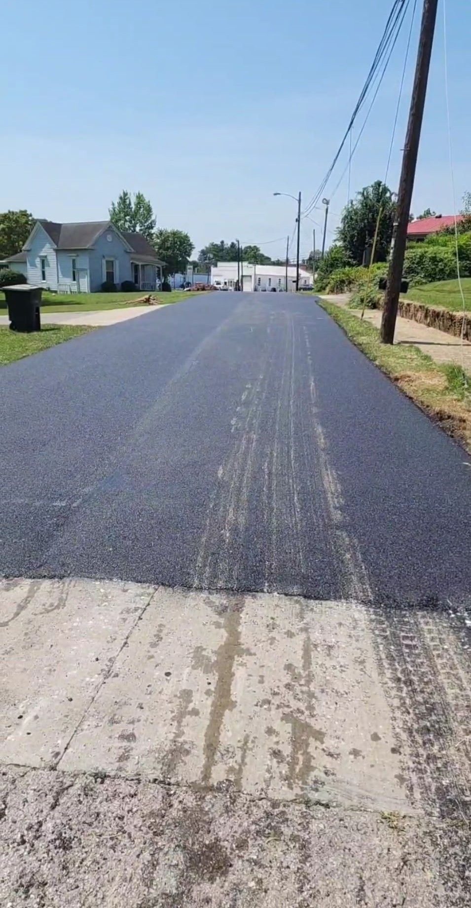 Freshly paved asphalt road with visible tire tracks, houses in the background, utility poles overhead