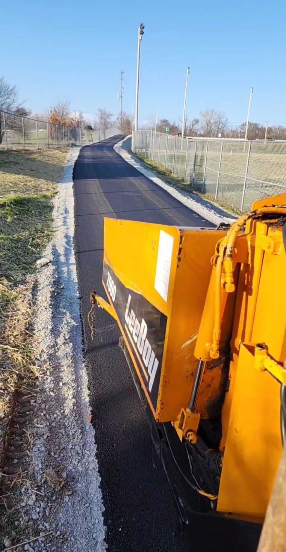 Asphalt paving machine laying fresh blacktop on a winding road next to grass and a chain-link fence