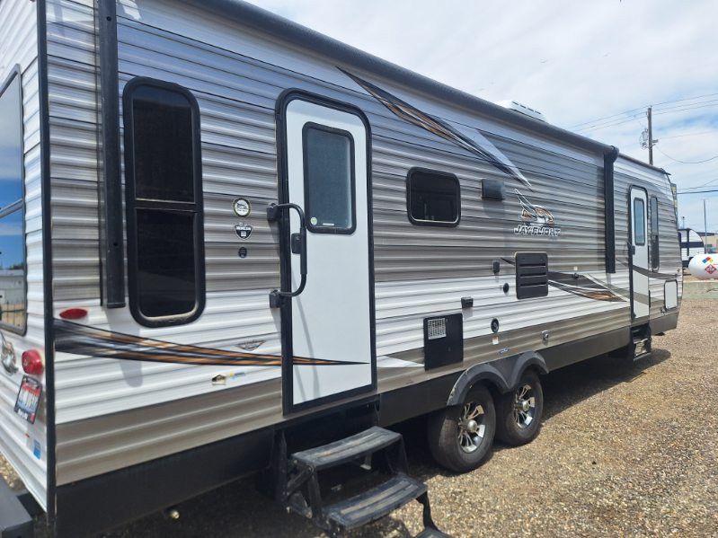 Silver and black travel trailer with side door and two axles parked on gravel under a cloudy sky