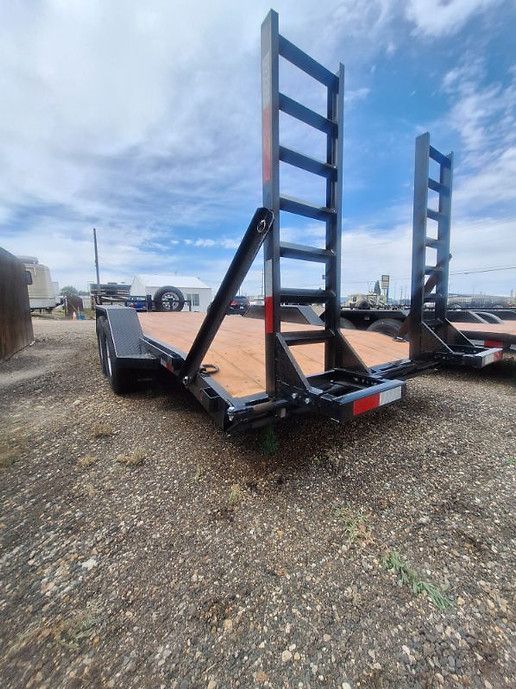 Flatbed trailer with tall black loading ramps parked on gravel in an open lot.