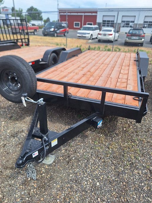 Flatbed utility trailer with black frame and orange wood deck parked on gravel.