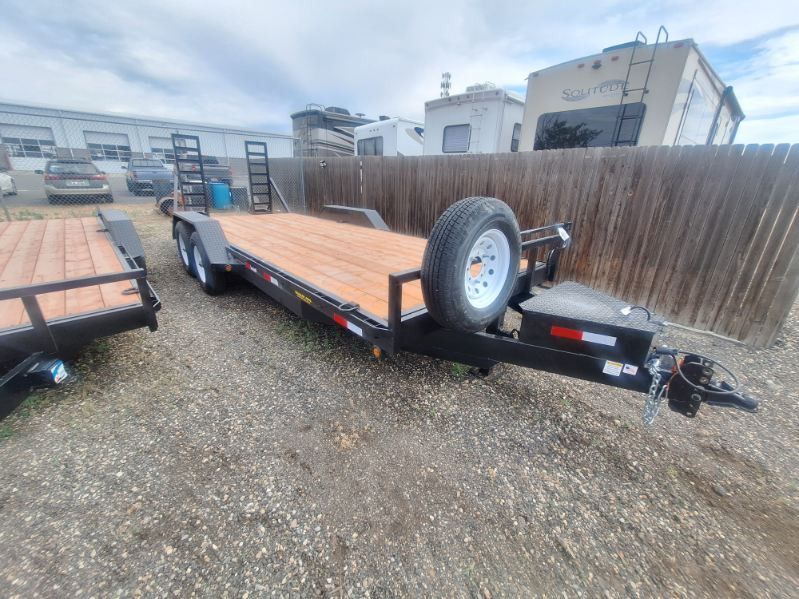 Flatbed utility trailer with wooden deck and spare tire, parked on gravel beside a wooden fence.