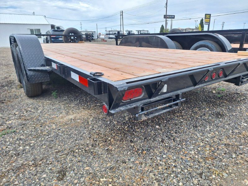 Black utility trailer with wooden deck parked on gravel lot under cloudy sky