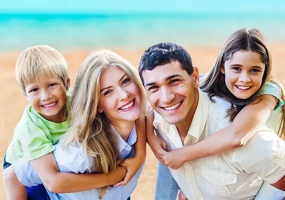 Family smiling at the beach, parents giving children piggyback rides. Turquoise water and blue sky.
