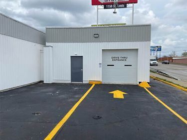 Drive-thru entrance at a car wash, with yellow directional arrows on black pavement, a garage door, and a gray door.
