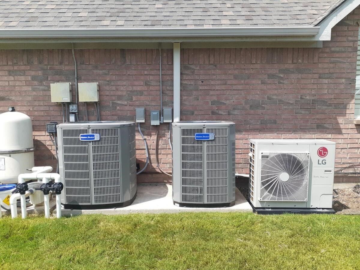 Three air conditioning units outside a brick building. Two are gray, one is white, all are next to a green lawn.