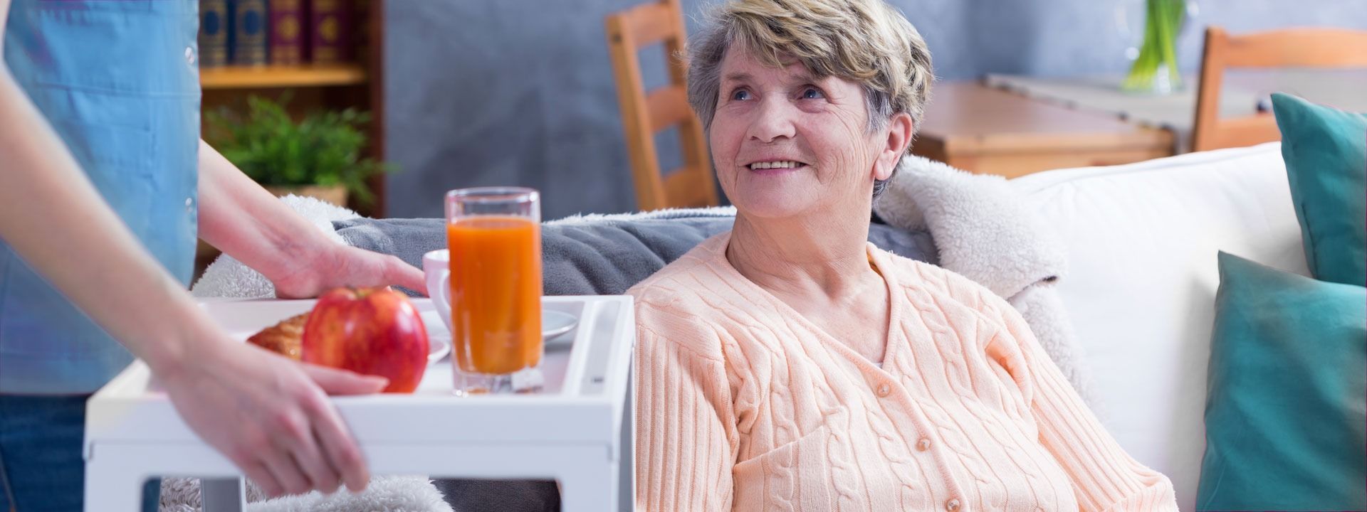 Caregiver serving a tray with orange juice to a smiling woman sitting on a couch