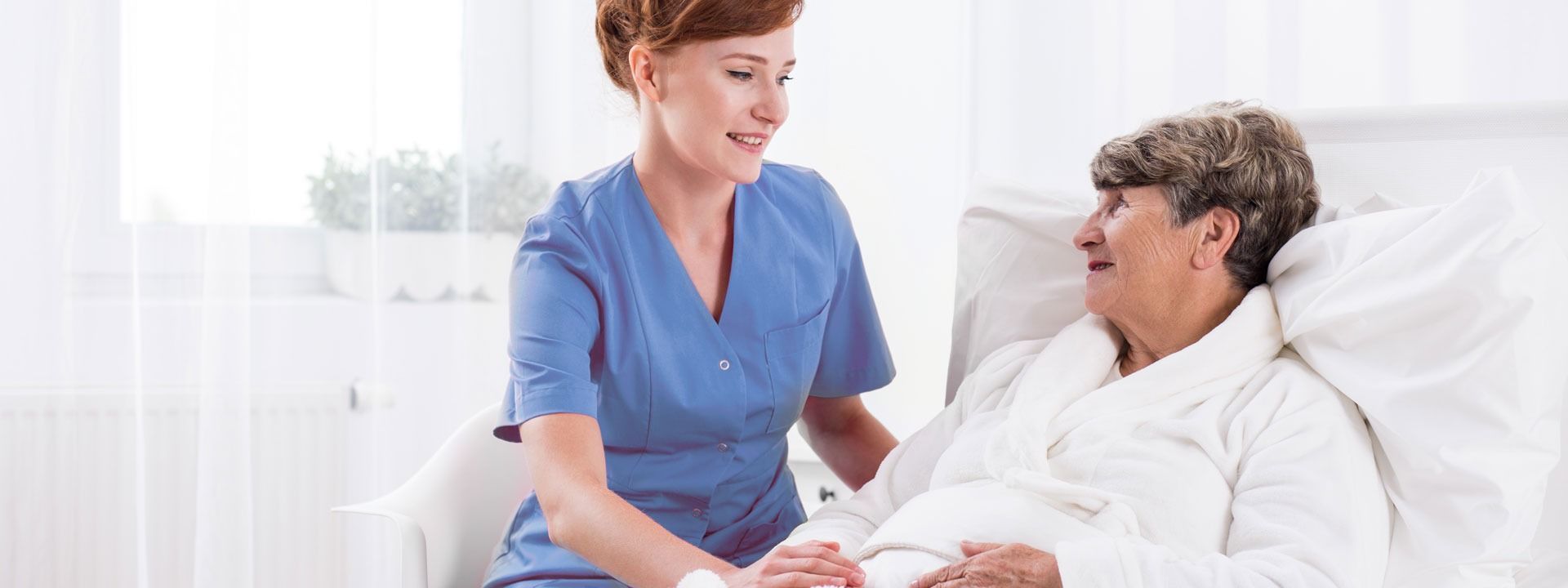 Nurse in blue scrubs talking to a patient in bed in a bright hospital room