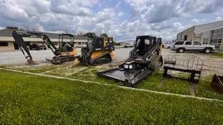 Construction equipment parked on grass: mini excavator, skid steer, and other attachments.