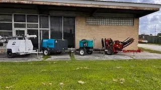 Construction equipment lined up on grass near a building; includes light tower, generators, and trencher.