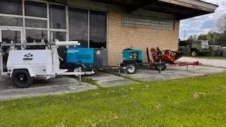 A white truck with equipment and other machinery sits outside a building with windows and green grass.