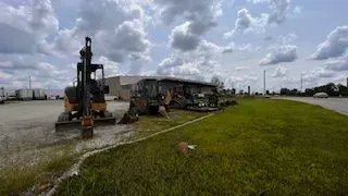 An excavator next to a damaged bus on a grassy area in front of a building on a cloudy day.