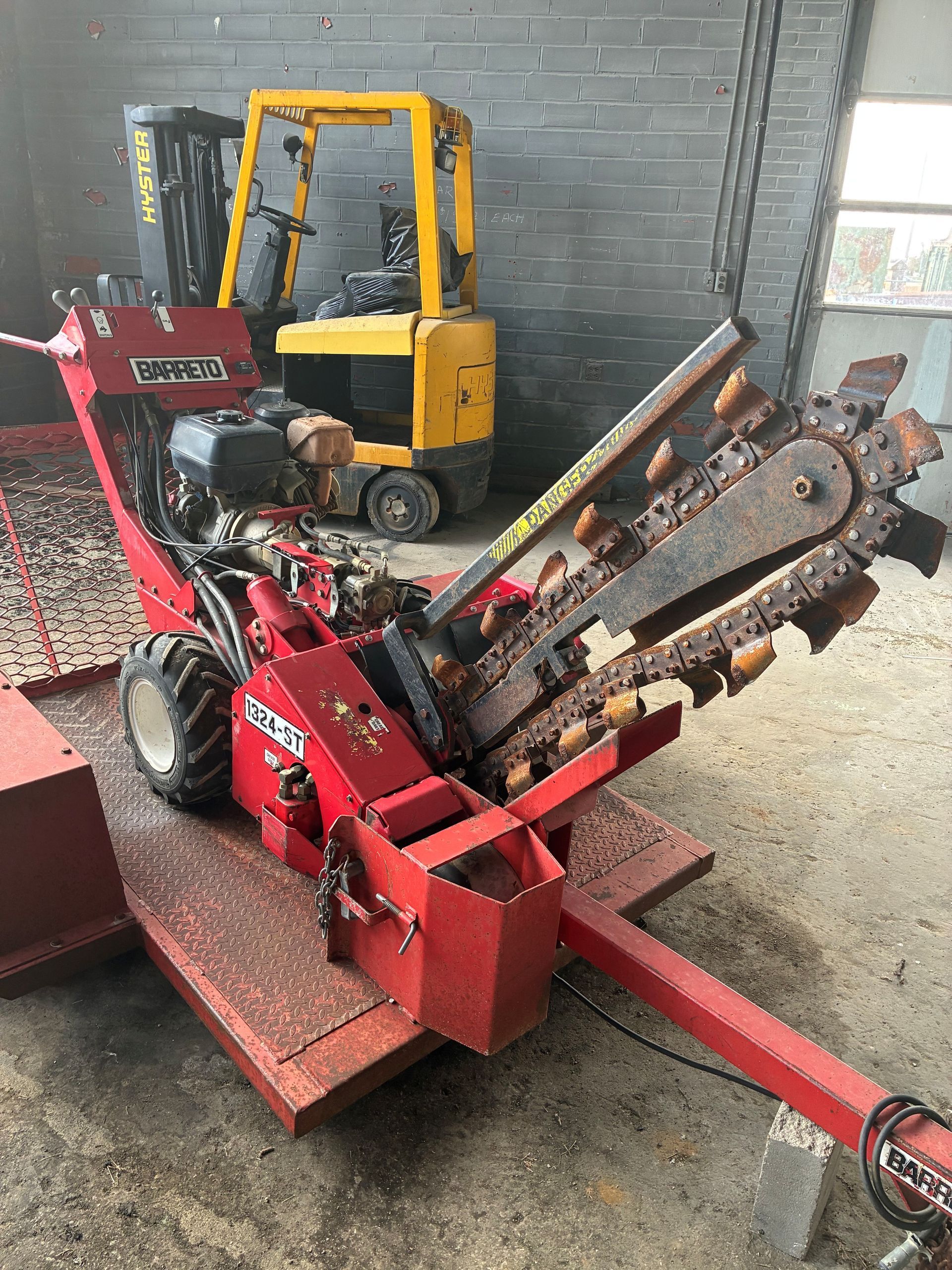 Red Ditch Witch trencher with cutting chain, parked inside; yellow forklift in the background.