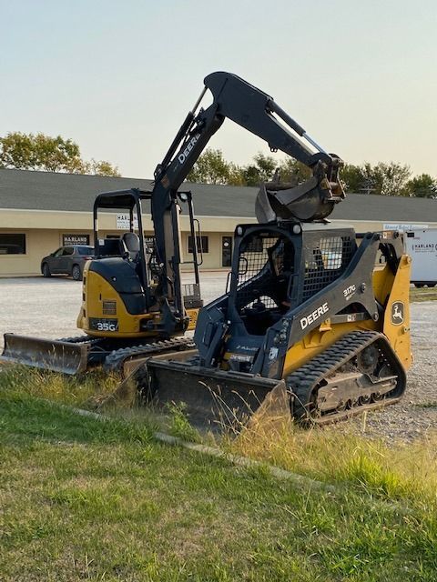 John Deere mini excavator and skid steer loader parked on grass in front of a building.