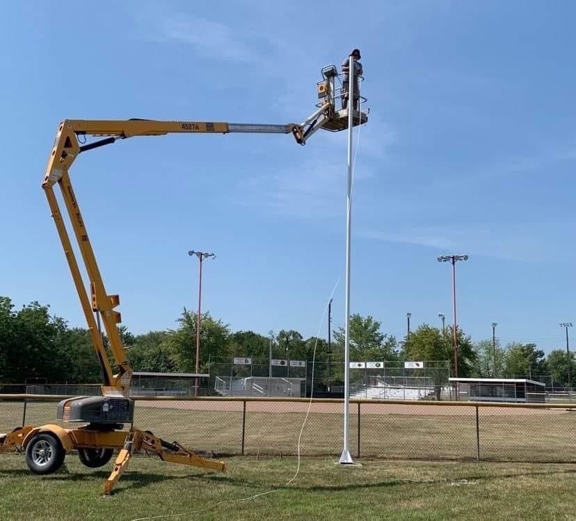 Yellow lift truck near a white flagpole on a baseball field, blue sky in background.