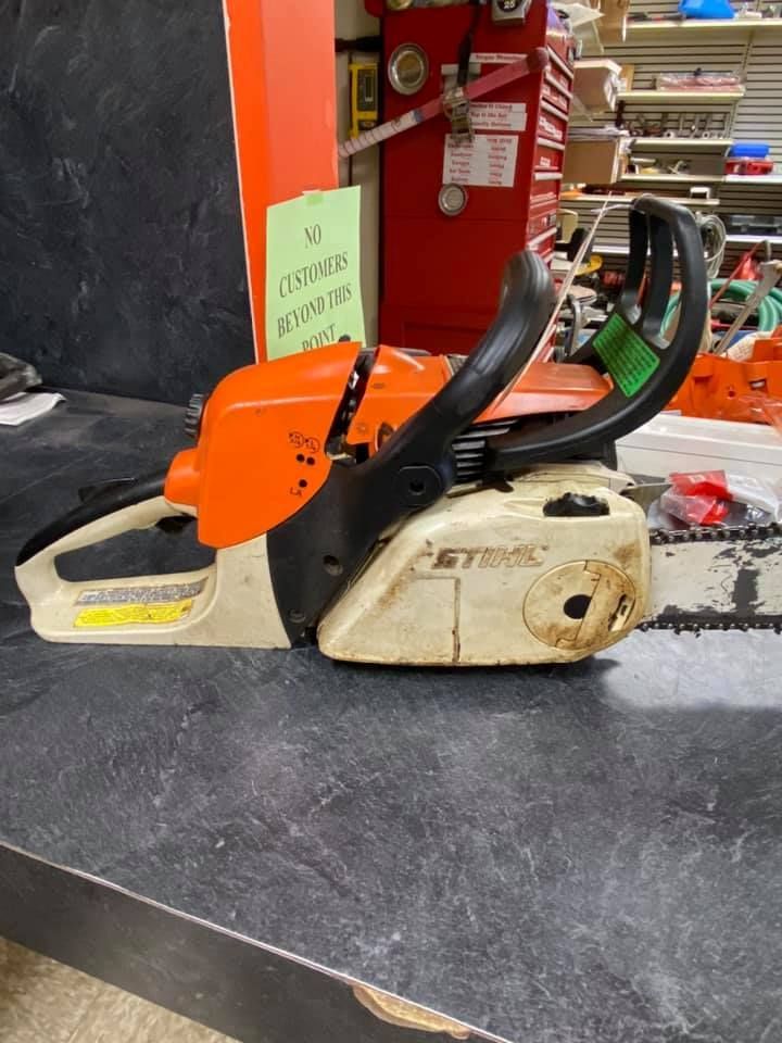 Stihl chainsaw on a black table. Orange and white body, black handle, in a shop.