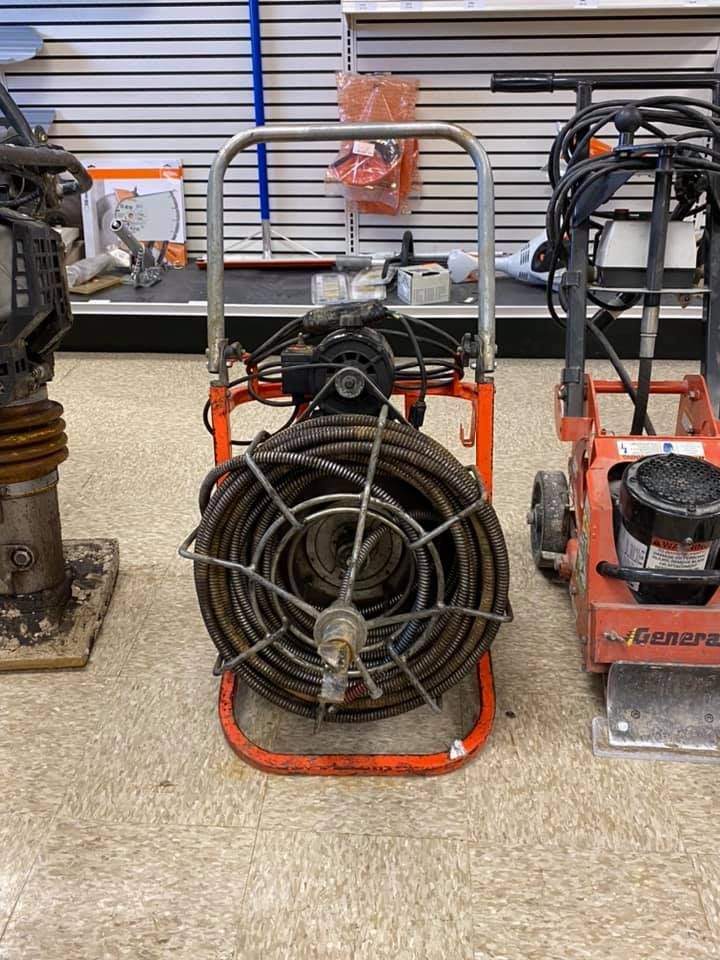 Orange plumbing snake machine with coiled cable, between a compactor and a tamper, indoors.