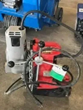 A red and gray magnetic drill press with a black handle, a black hose, and green tag, on a concrete floor.