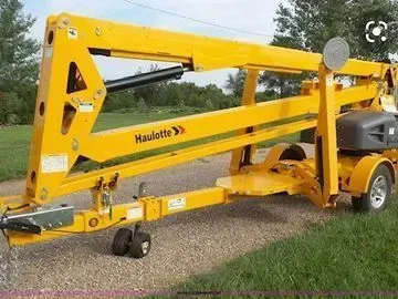 Yellow Haulotte trailer-mounted boom lift parked on grass; for raising workers to reach high places.