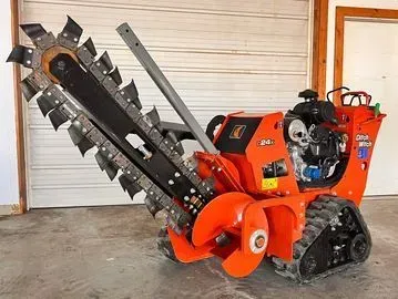 Orange trenching machine with rotating blade and tracks, near a garage door.
