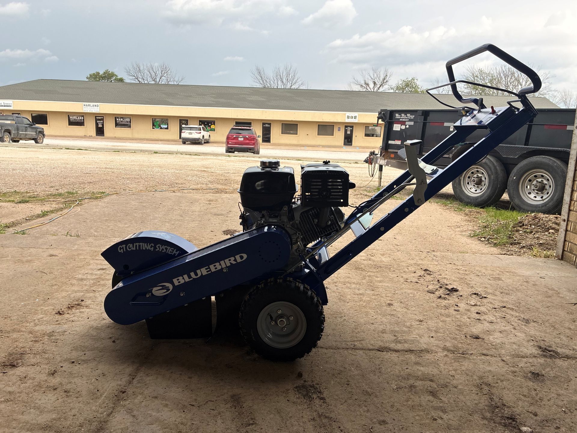 Blue wood chipper parked on gravel beside a building, with a truck in the background.