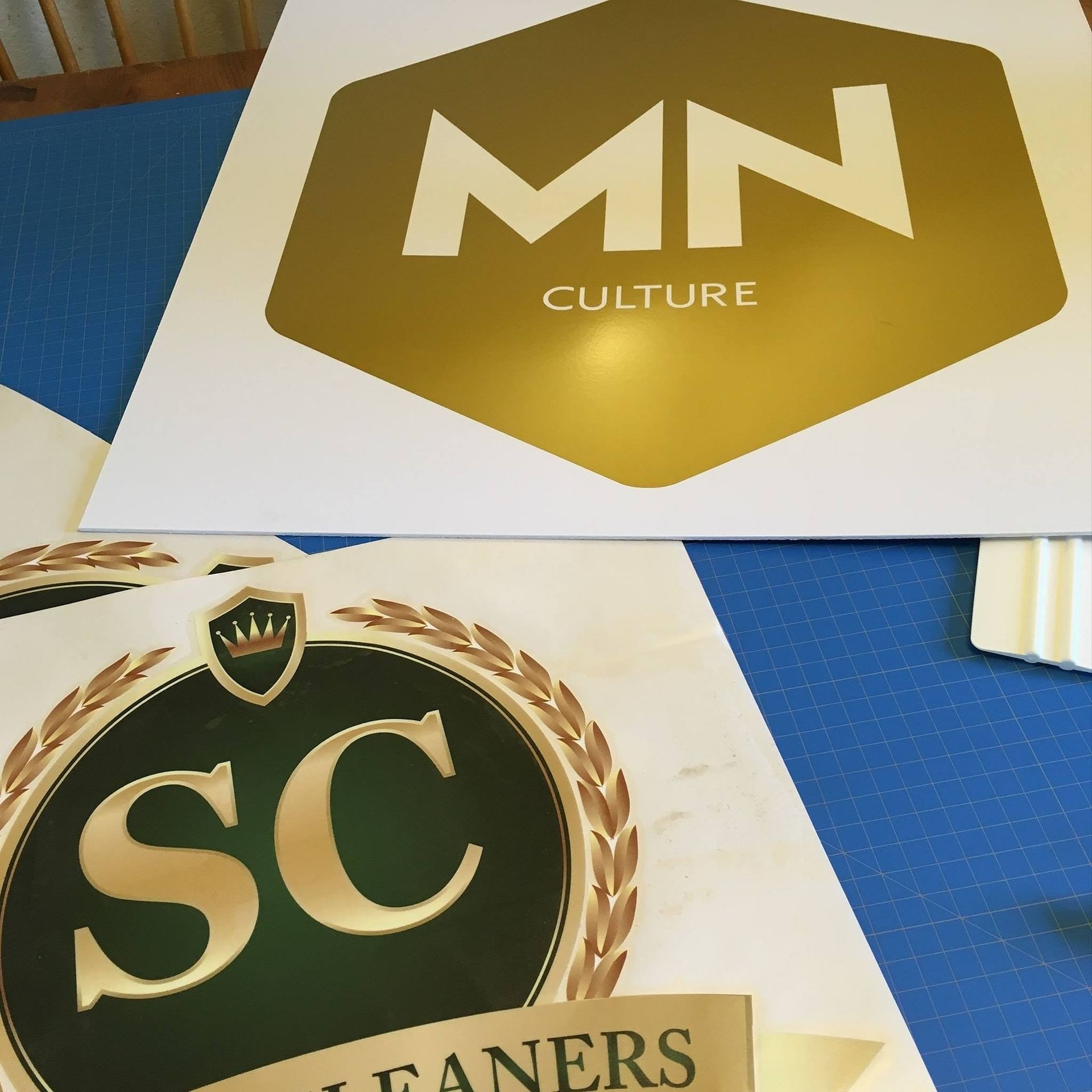 Two gold-and-black logo cards on a blue surface, reading “MN Culture” and “SC Saners.”