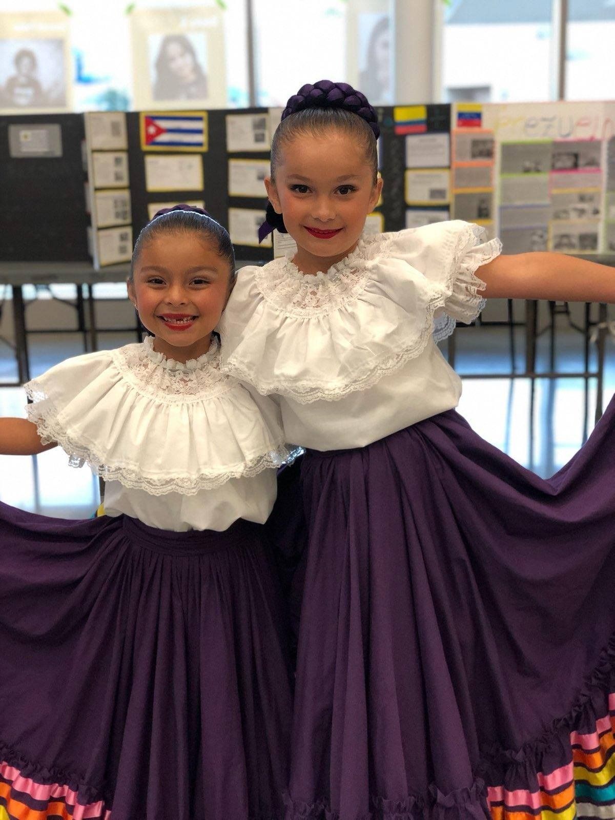 Two children in white ruffled tops and purple skirts smiling indoors, arms raised together.