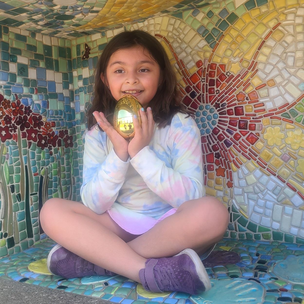 Person sitting cross-legged in a mosaic alcove, holding a glowing object and smiling
