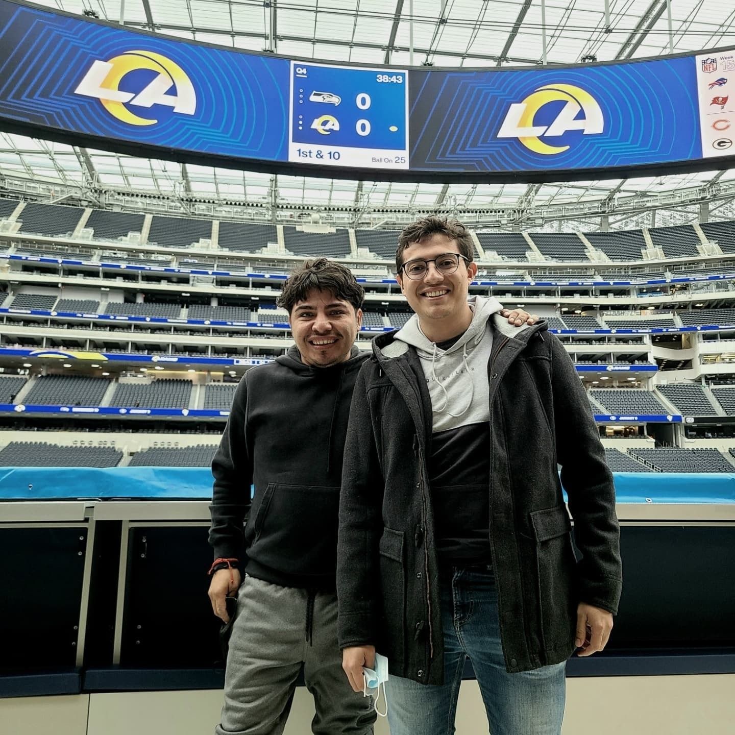 Two smiling people standing in an empty football stadium with LA Rams logos on the scoreboard