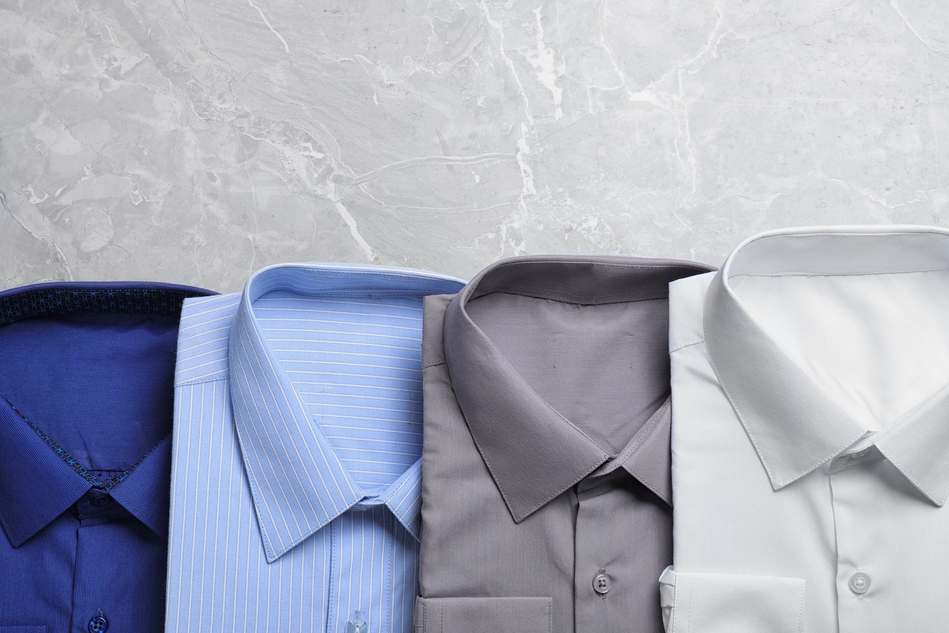 Four folded dress shirts in blue, light blue, gray, and white on a textured gray background
