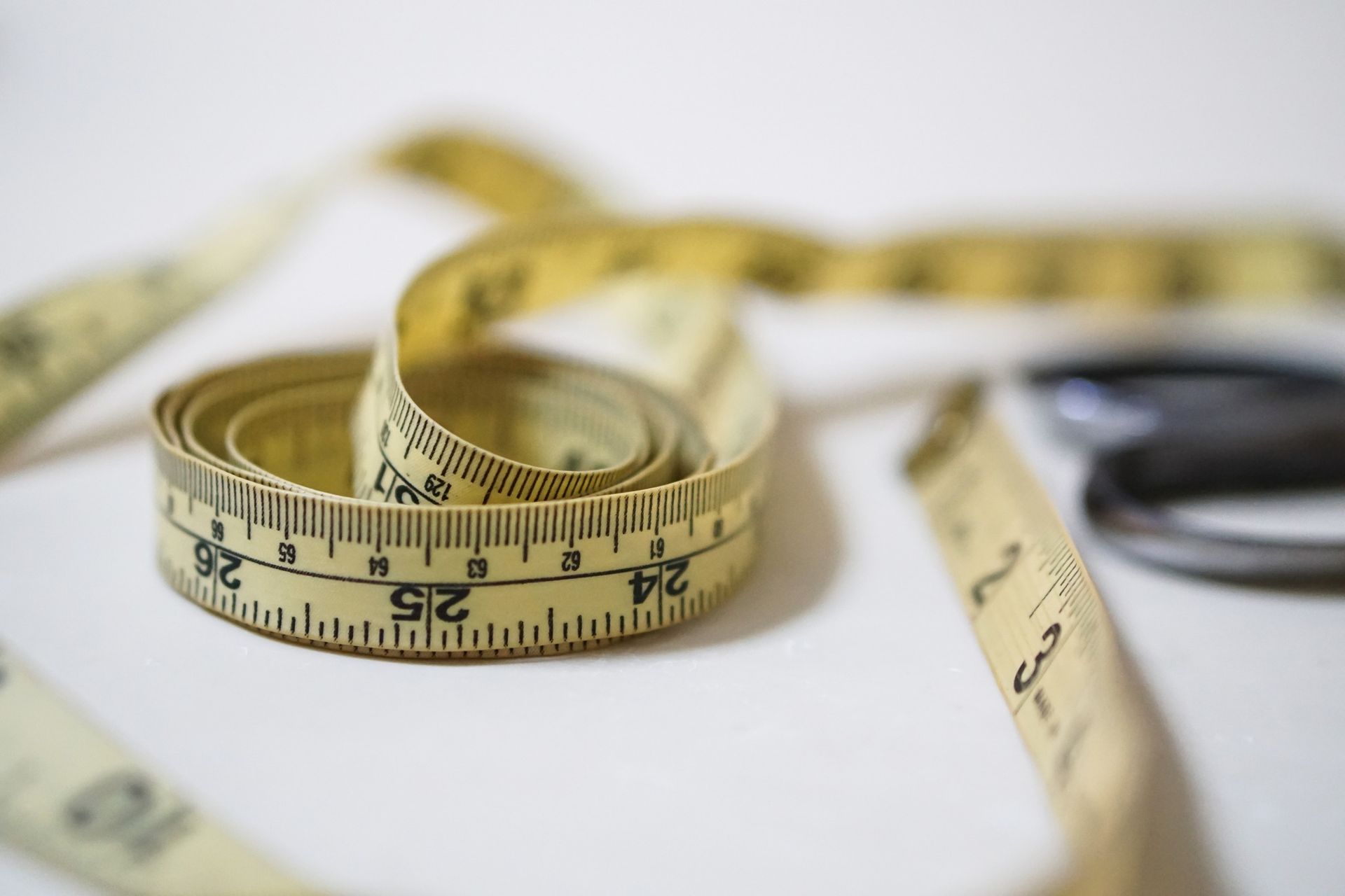 Coiled yellow measuring tape on a white surface, with blurred black scissors in the background