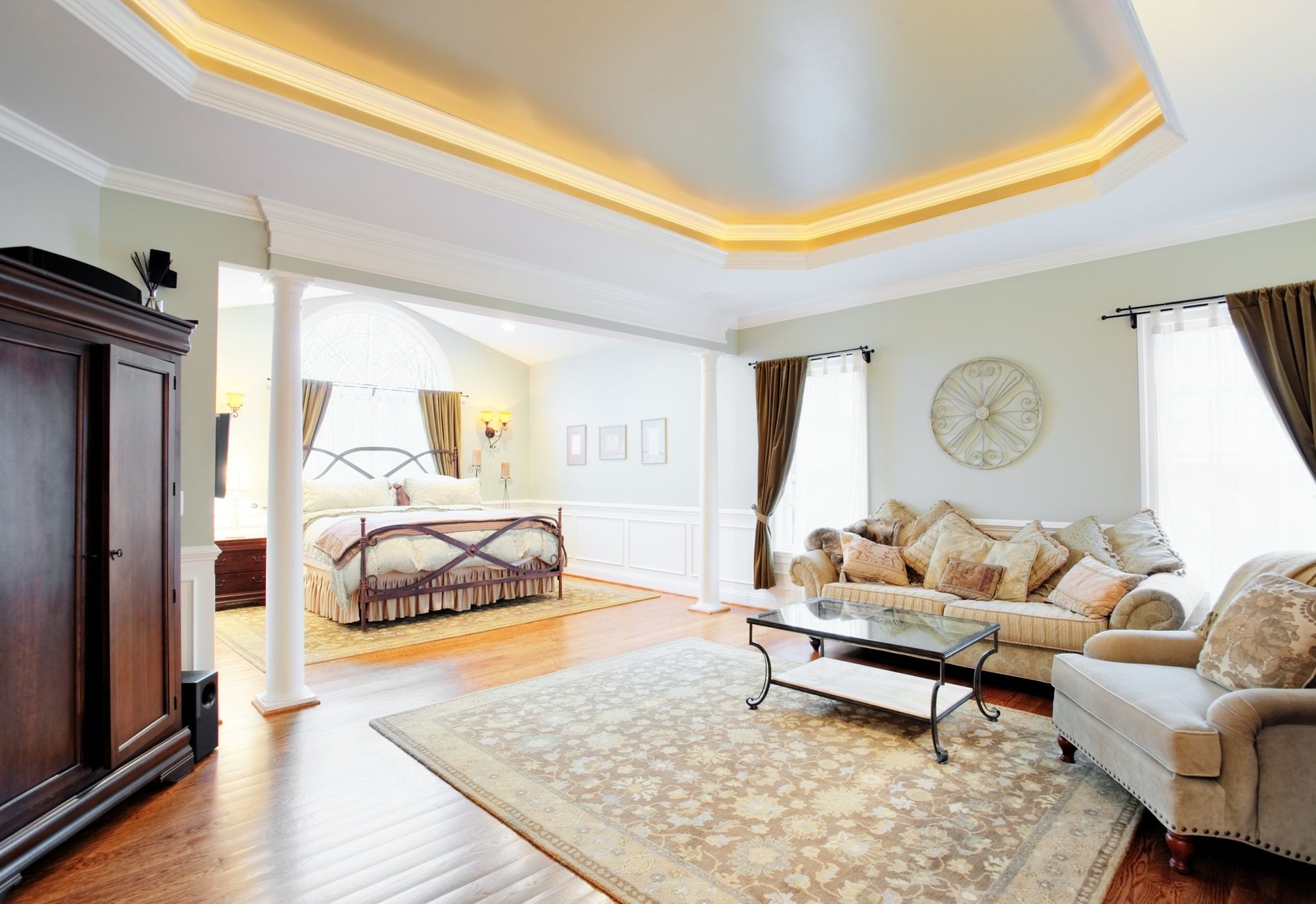 Bright living room with beige sofas, patterned rug, wall clock, and adjacent bedroom visible through an archway