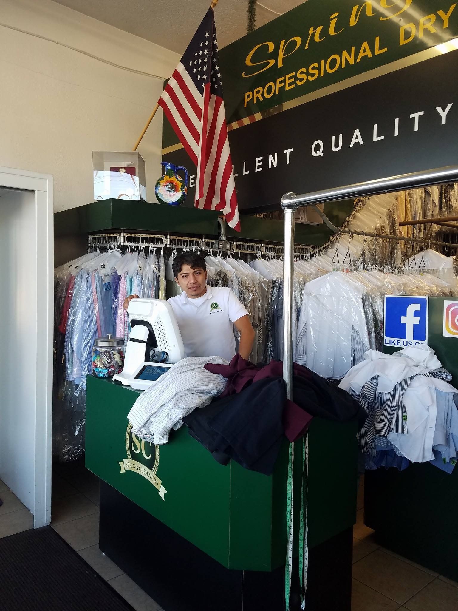 Dry cleaner counter with hanging shirts, folded clothes, American flag, and visible social media signs