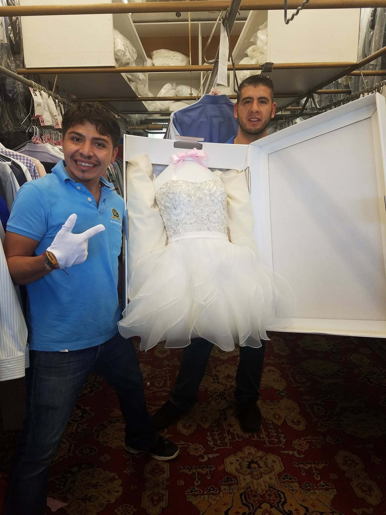 Two men holding up a white tulle dress in a clothing shop, one smiling and pointing at it