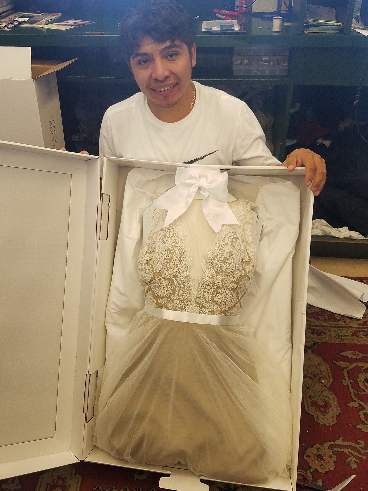 Person holding a boxed formal white gown with a large bow and embroidered bodice indoors