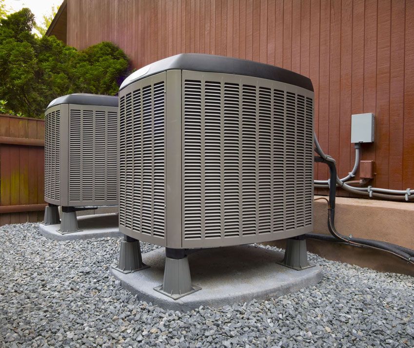 Two air conditioners are sitting on gravel outside of a house.