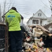 Man in green hoodie next to a dumpster filled with trash, in front of houses.