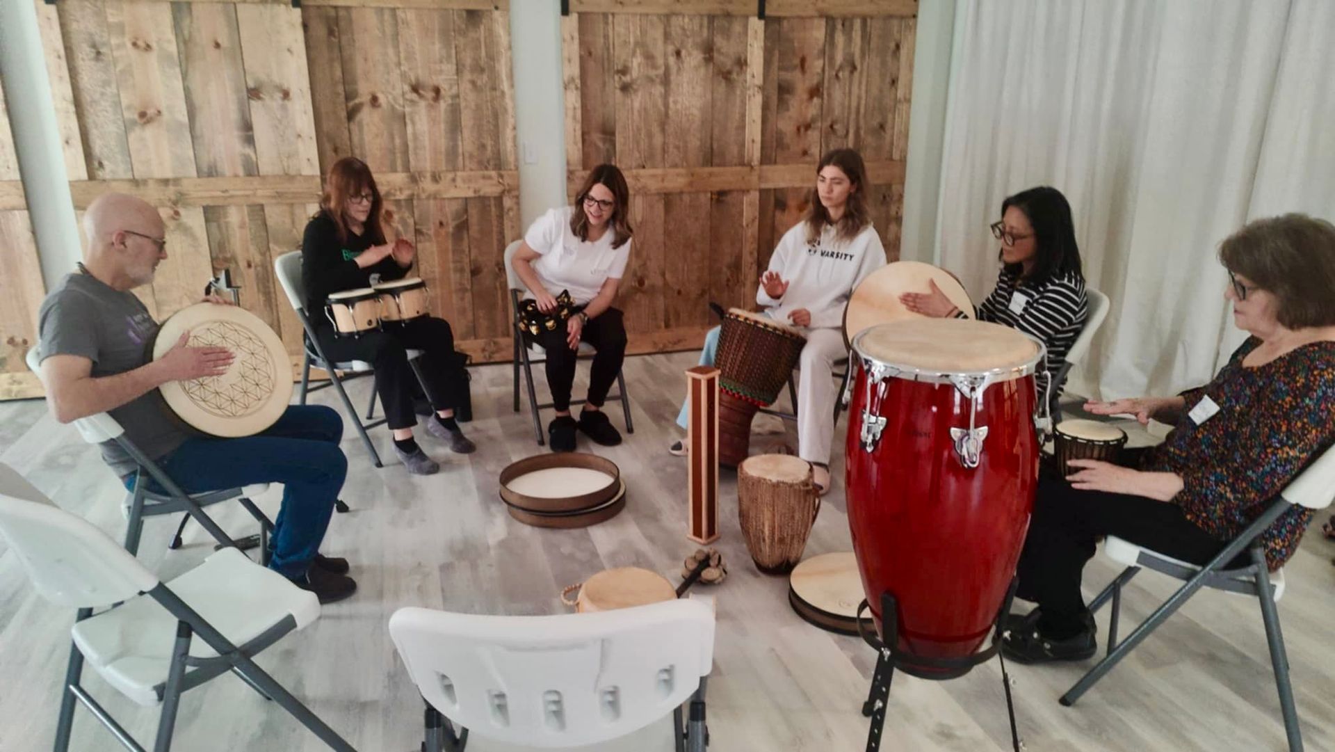 Group of people sitting in a circle playing various drums in a room with wood paneling.