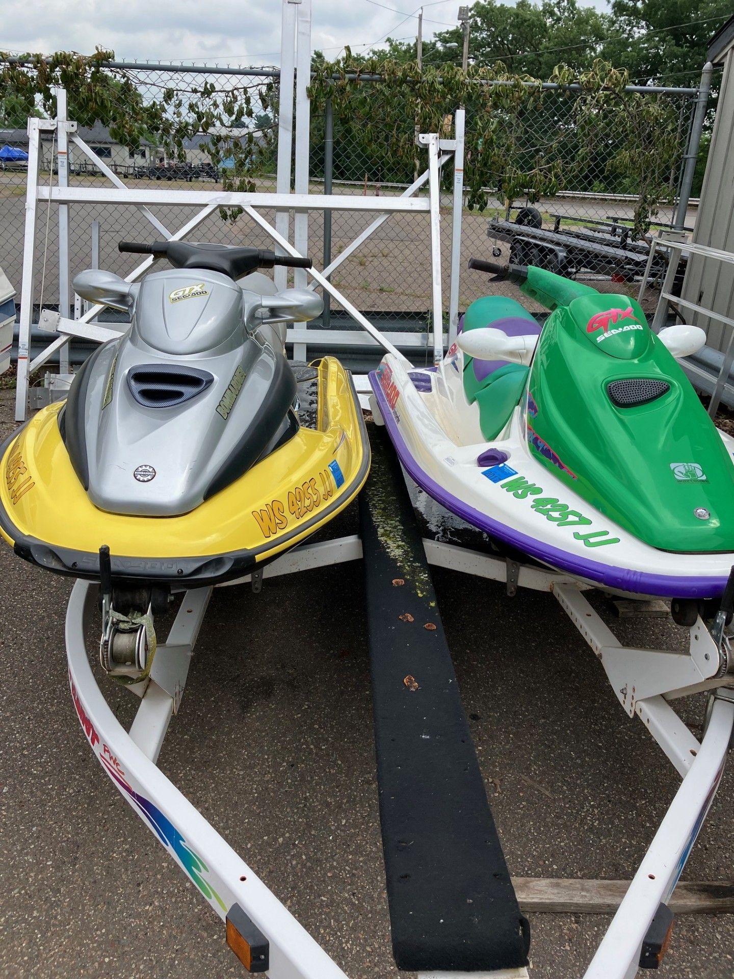 Two personal watercraft, one silver and yellow and one green and white, sit side-by-side on a boat trailer. 